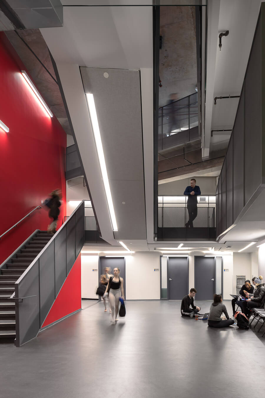 Hallway of the School of Performance at Ryerson University. A double-height red wall juxtaposes against warm, light and dark greys on the polished concrete floors and inter-secting stairwells. The space looks up from the lower level to the mezzanine and the open stairs going to the upper levels. Strips of warm, bright lighting illuminate the space as dancers prepare for classes.