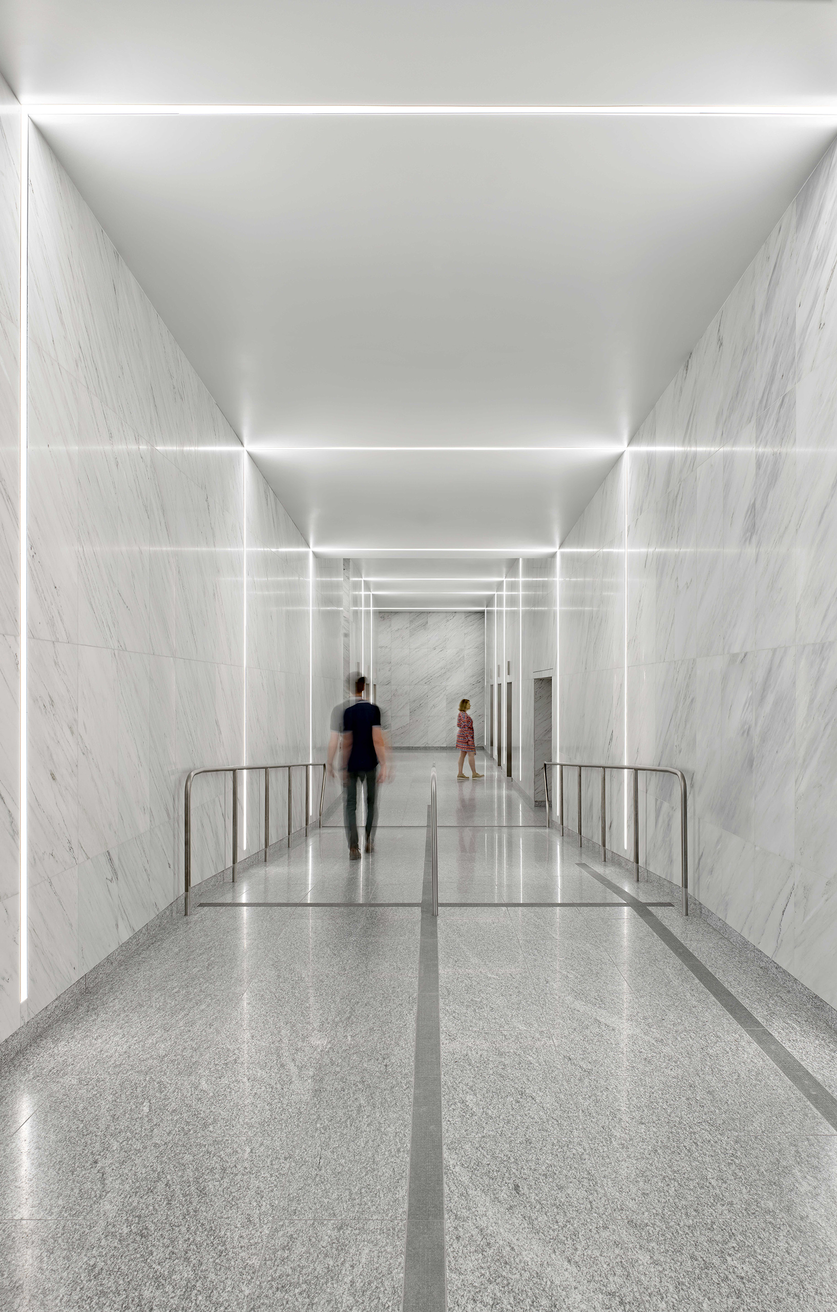 Interior lights illuminate the corridor, where two individuals are walking away from the main perspective. The white marble walls are illuminated by the lighting, creating a bright white interior space.